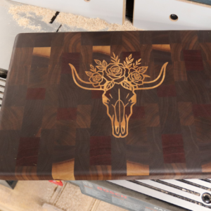 End grain cutting board with walnut and mahogany checker base and a maple inlay of a steer skull crowned with roses — handmade by Langskegg